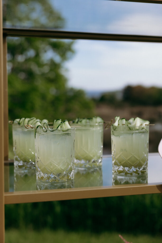 Vermont Wedding Venues | Sincerely Liz Photography Documentary and Editorial Wedding Photography | Five glasses of a pale green cocktail garnished with cucumber ribbons sit on a glass shelf, with a blurred background of lush greenery and blue sky.