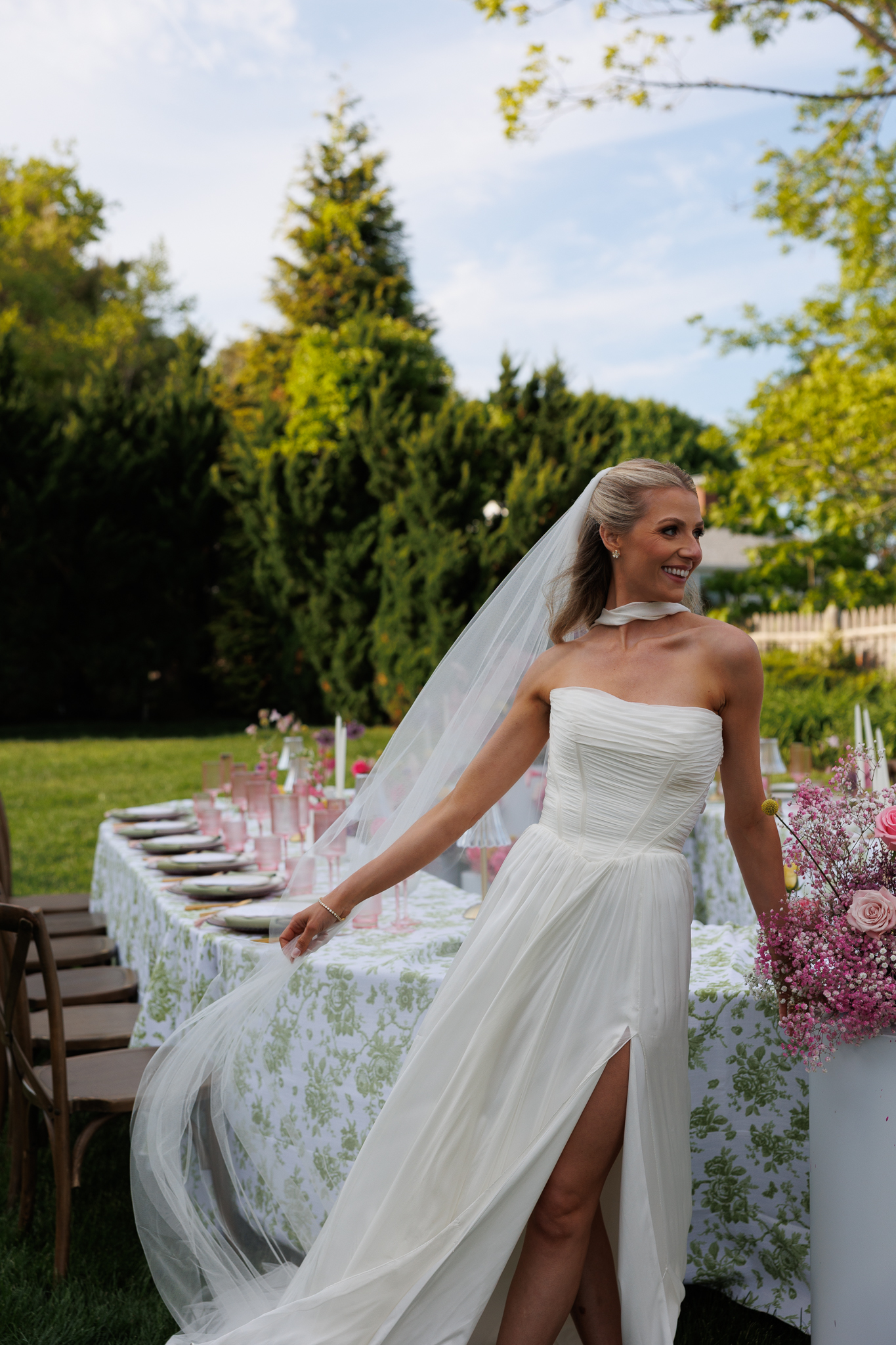 Vermont Wedding Venues | Sincerely Liz Photography Documentary and Editorial Wedding Photography | A bride in a white dress with a high slit and veil stands by a floral-decorated outdoor table, surrounded by greenery, looking serene and joyful.