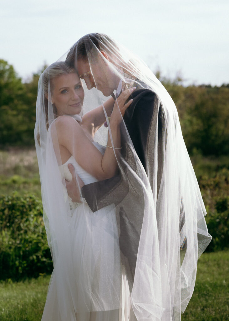 Vermont Wedding Photographer | Sincerely Liz Photography Documentary and Editorial Wedding Photography | Bride and groom in an intimate embrace under a flowing veil, standing outdoors surrounded by greenery. They appear joyful and serene.
