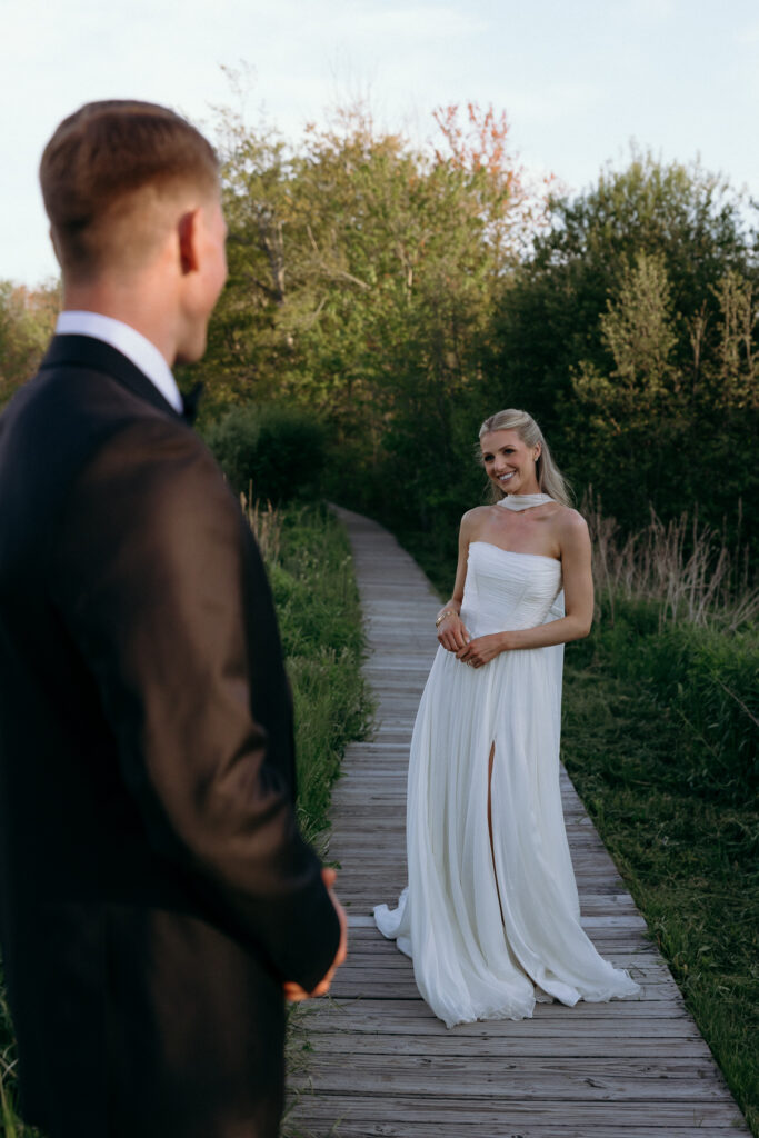 Groom watches as the bride stands at the end of a wooden path in golden evening light, a romantic frame that fits wedding venues for 50 guests Vermont couples consider.