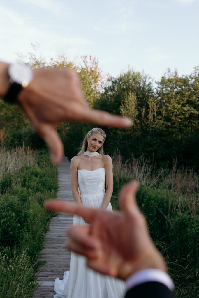 Wedding Photographers in Vermont | Sincerely Liz Photography Documentary and Editorial Wedding Photography | Bride in a white dress stands on a boardwalk surrounded by greenery. Framing hands in the foreground suggest a playful, candid moment.