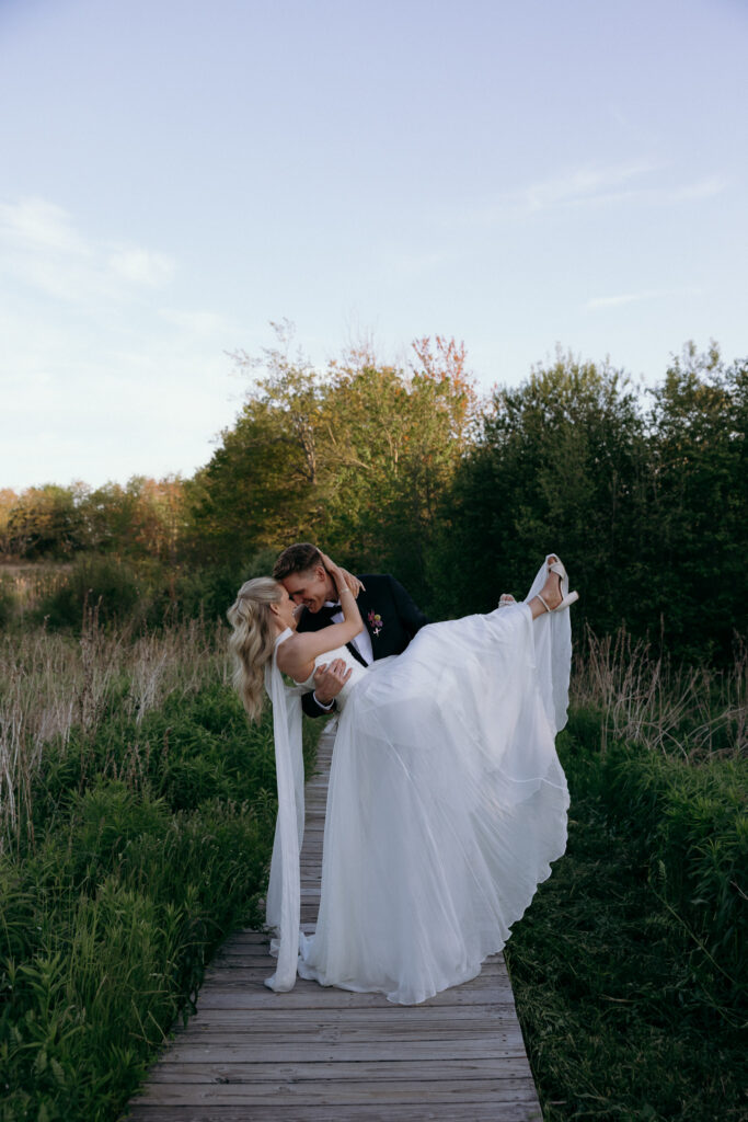 Vermont Wedding Photographer | Sincerely Liz Photography Documentary and Editorial Wedding Photography | A man in a suit joyfully lifts and kisses a woman in a flowing white gown on a wooden path through a lush, green field under a clear blue sky.