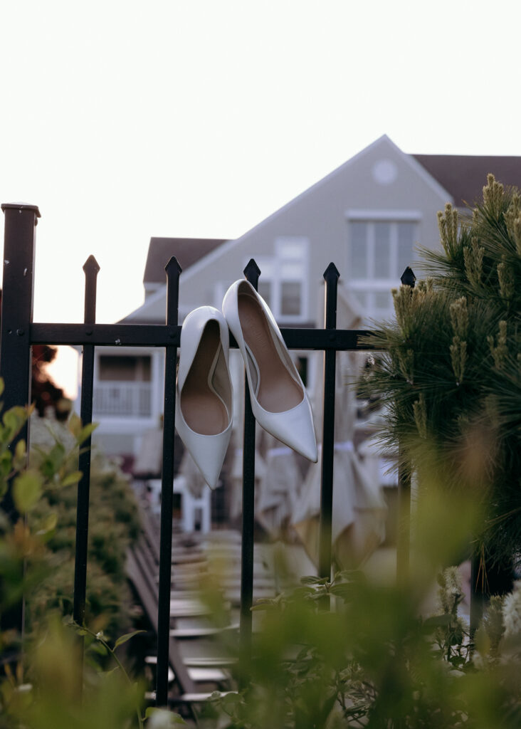 Bridal shoes hang from a black iron fence with a white venue building softly blurred in the background, an elegant detail from wedding venues in VT.