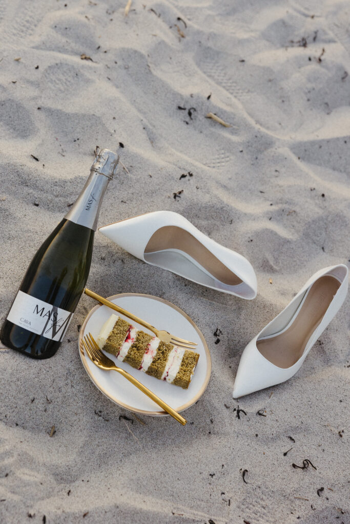 Wedding Venue Vermont | Sincerely Liz Photography Documentary and Editorial Wedding Photography | White high heels, a bottle of sparkling wine, and a slice of cake with gold utensils on a plate are artfully arranged on sandy beach.