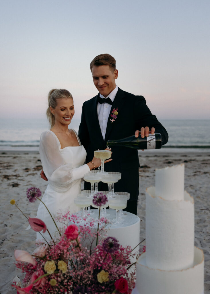 Wedding Photographers in Vermont | Sincerely Liz Photography Documentary and Editorial Wedding Photography | A couple toasts on the beach at sunset. The man pours champagne into a tower of glasses. Bright flowers and a cake are in the foreground. Romantic and joyful atmosphere.