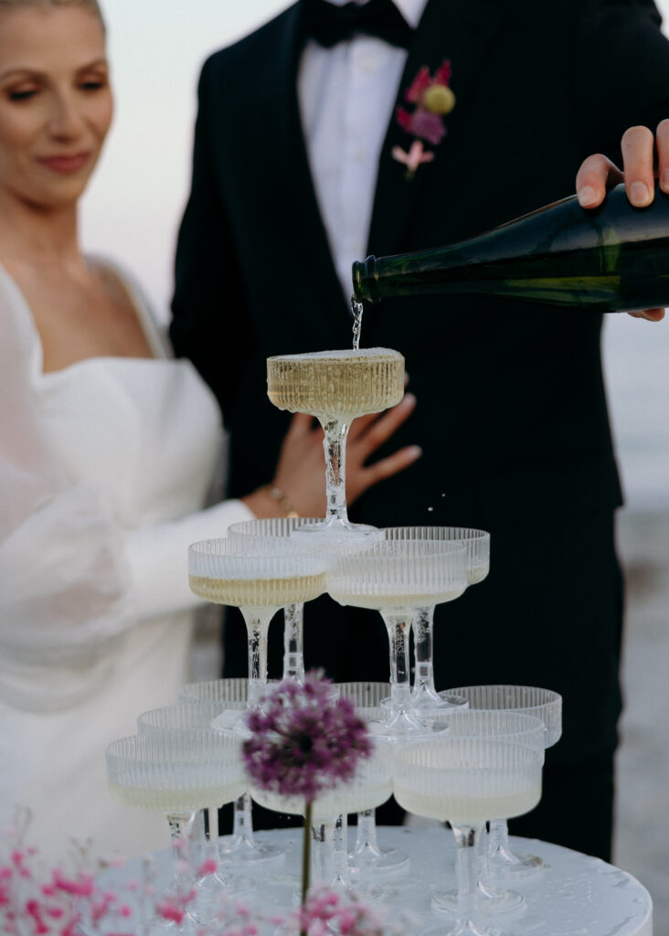 Vermont Wedding Venues | Sincerely Liz Photography Documentary and Editorial Wedding Photography | A bride in a white dress and groom in a suit pour champagne into a tower of glass coupes. The setting is elegant, with delicate flowers nearby.