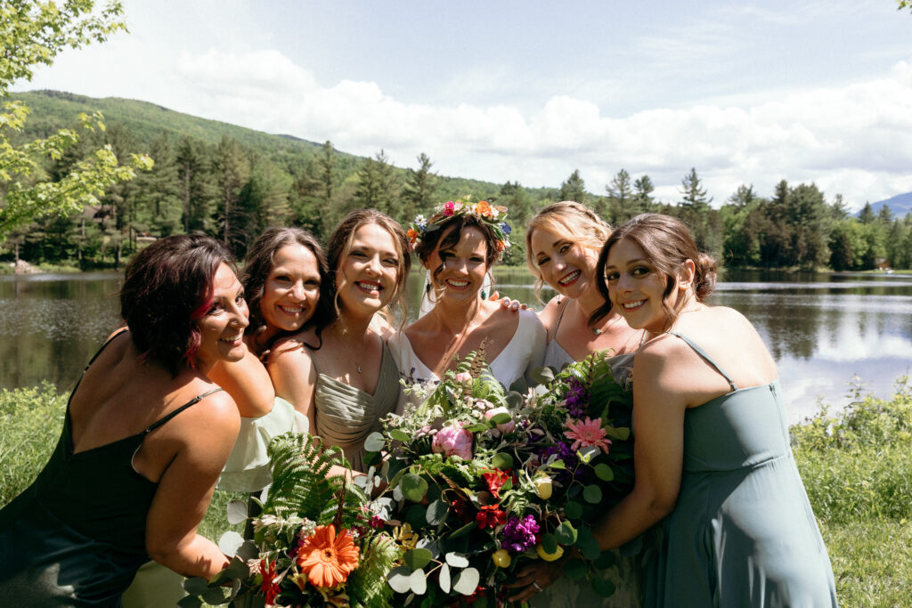 Bride and bridesmaids gather beside a peaceful lake framed by mountains and wild greenery, showcasing the appeal of lakeside wedding venues in Vermont and destination celebrations surrounded by nature.