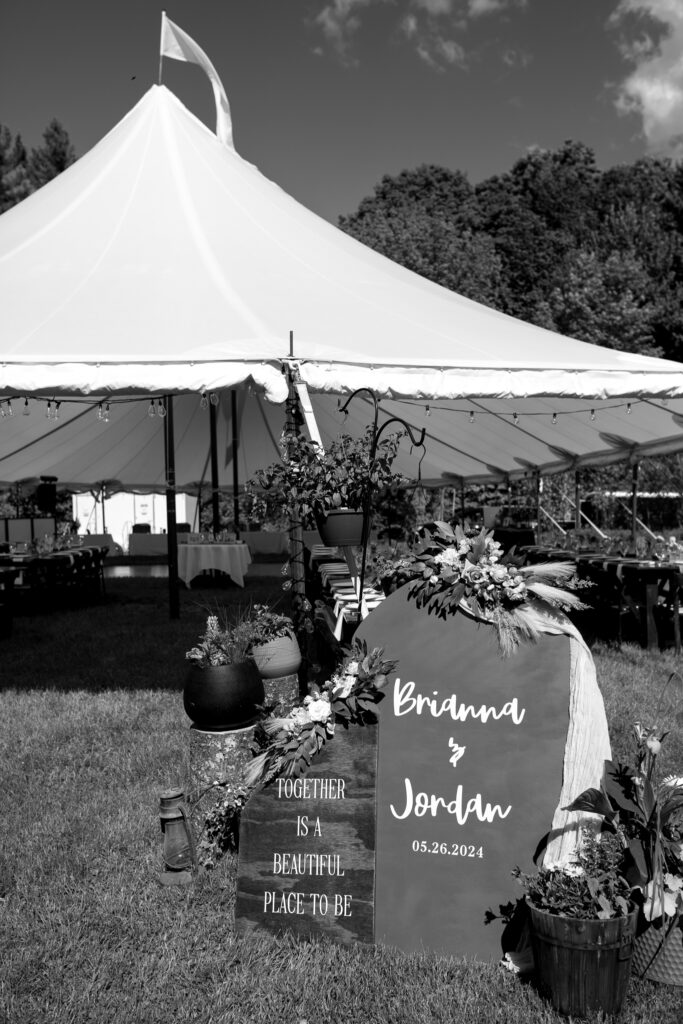 Vermont Wedding Venues | Sincerely Liz Photography Documentary and Editorial Wedding Photography | Black and white image of an outdoor wedding tent with a sign displaying "Brianna & Jordan, 05.26.2024." Decor includes flowers and rustic lanterns, conveying a romantic and celebratory atmosphere.