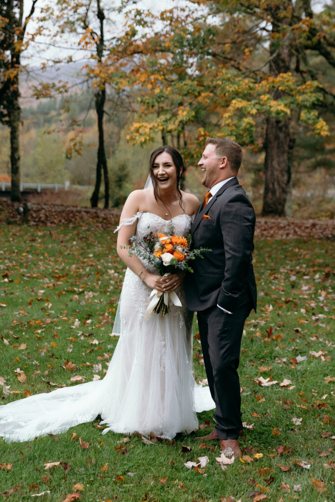 Vermont Wedding Venues | Sincerely Liz Photography Documentary and Editorial Wedding Photography | A bride in a white lace dress holds orange and white flowers, standing with a groom in a dark suit. Both are smiling in a leafy autumn park.
