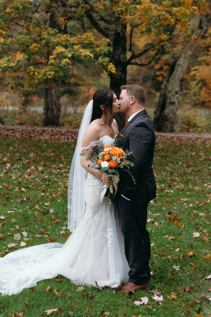Vermont Wedding Photographer | Sincerely Liz Photography Documentary and Editorial Wedding Photography | A bride and groom kiss outdoors on a grassy lawn with autumn leaves. The bride wears a white dress and veil, holding a bouquet of orange flowers.