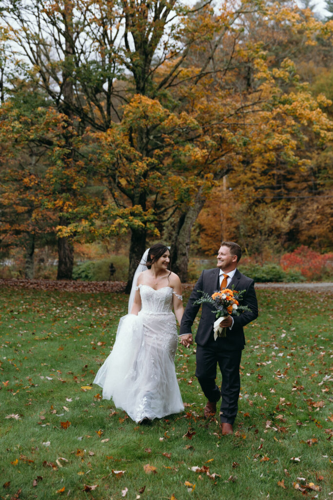 Vermont Wedding Photographer | Sincerely Liz Photography Documentary and Editorial Wedding Photography | A bride in a white gown and groom in a dark suit walk hand in hand across a grassy field, surrounded by autumn trees and falling leaves, exuding joy.