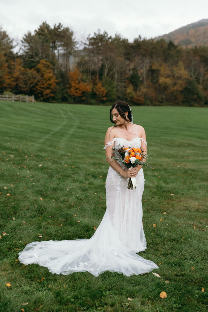 Vermont Wedding Venues | Sincerely Liz Photography Documentary and Editorial Wedding Photography | A bride in an off-shoulder white gown holds a vibrant bouquet with orange and white flowers, standing on green grass, with autumn trees in the background.