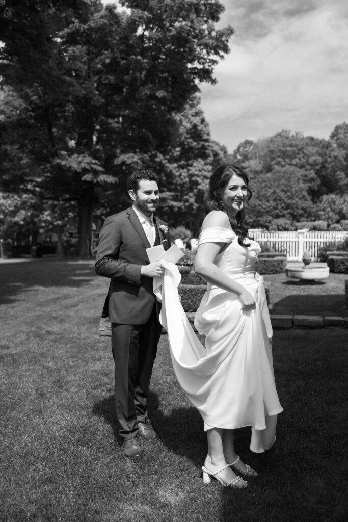 Outdoors on the lawn, the groom adjusts the bride’s dress as she smiles back at him, reflecting the relaxed and connected experience of a Smith Farm Gardens wedding.
