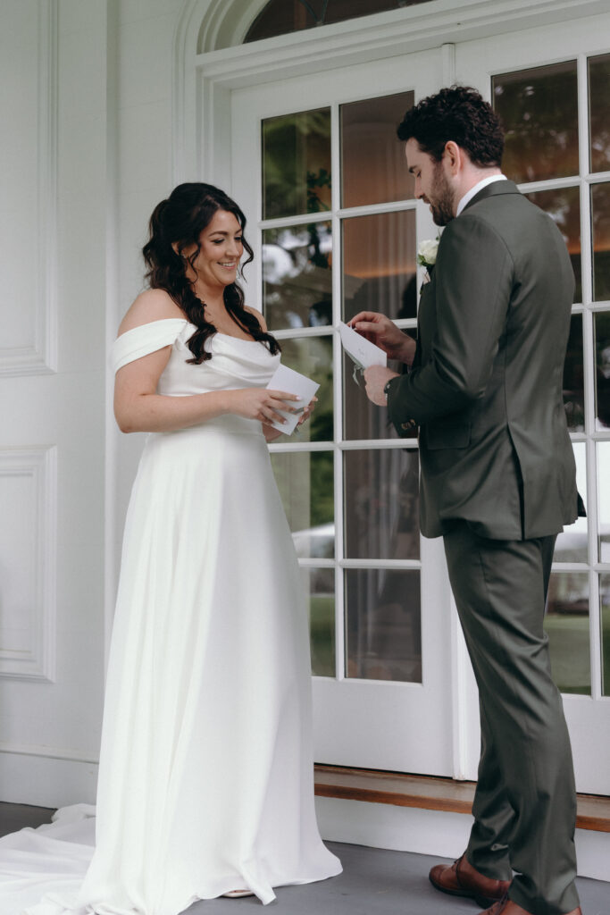 Smith Farm Gardens Wedding | Sincerely Liz Photography Documentary and Editorial Wedding Photography | A bride in an off-shoulder white dress and a groom in a dark suit stand at a doorway exchanging vows. Both smiling, creating a warm, joyful atmosphere.