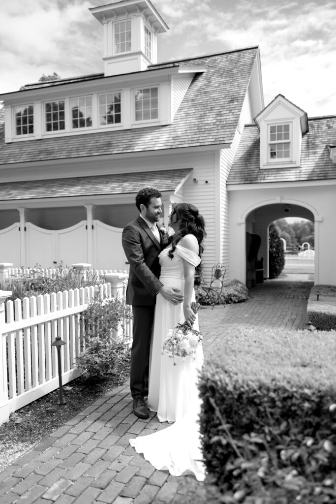 Smith Farm Gardens Wedding | Sincerely Liz Photography Documentary and Editorial Wedding Photography | A couple stands lovingly in front of a charming house with a white picket fence. The bride holds a bouquet, and both wear classic wedding attire.