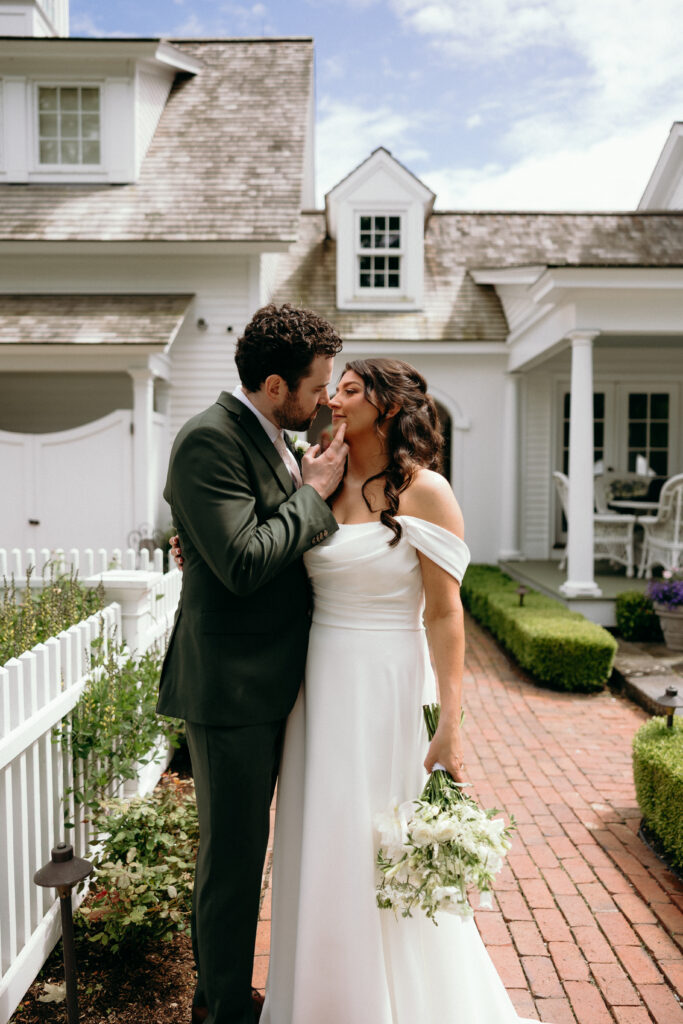 The bride and groom stand close along a brick pathway outside a coastal-style home, framed by manicured greenery that echoes timeless Vermont estate wedding venues.