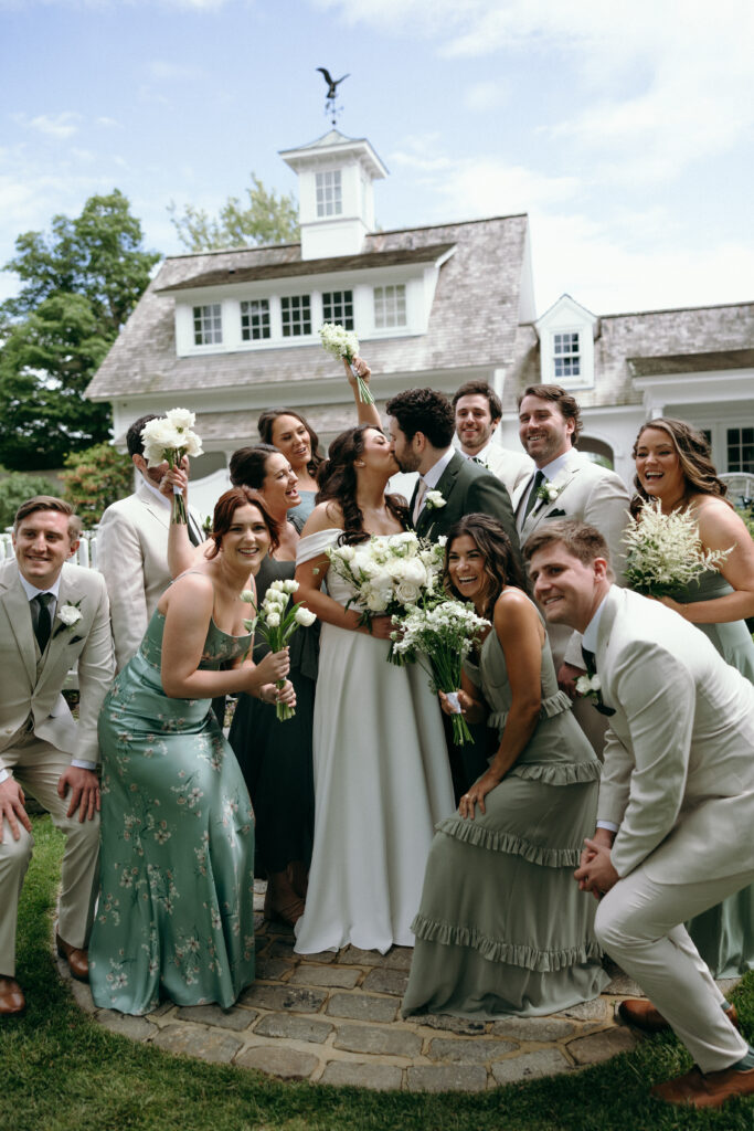 Smith Farm Gardens Wedding | Sincerely Liz Photography Documentary and Editorial Wedding Photography | A newlywed couple kisses, surrounded by a joyful wedding party in elegant attire. They stand outside a rustic house, conveying celebration and happiness.