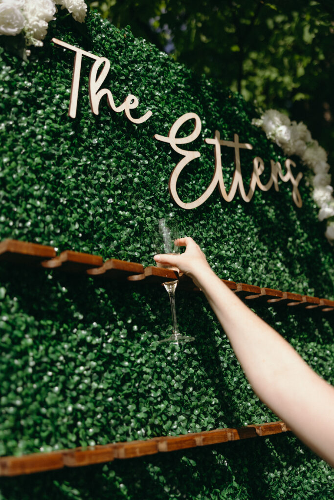 Smith Farm Gardens Wedding | Sincerely Liz Photography Documentary and Editorial Wedding Photography | A hand reaches to take a champagne flute from a decorative green wall with wooden holders. "The Etters" is written above in elegant script, with white flowers nearby.
