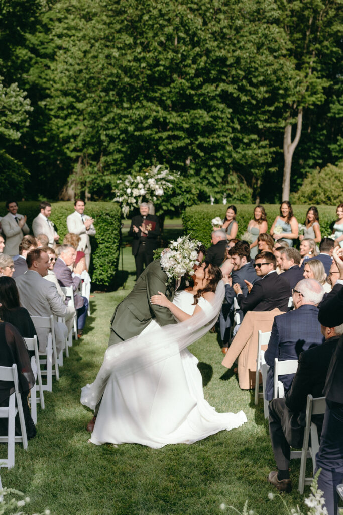 Smith Farm Gardens Wedding | Sincerely Liz Photography Documentary and Editorial Wedding Photography | A joyful wedding scene with a couple kissing passionately at the aisle's center, surrounded by smiling guests seated outdoors, lush greenery, and sunny weather.