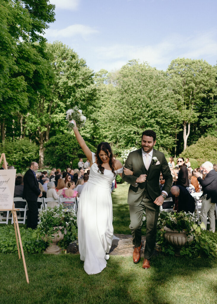 Smith Farm Gardens Wedding | Sincerely Liz Photography Documentary and Editorial Wedding Photography | Bride in a white dress and groom in a green suit joyfully exit an outdoor ceremony, surrounded by cheering guests and lush greenery under a clear sky.