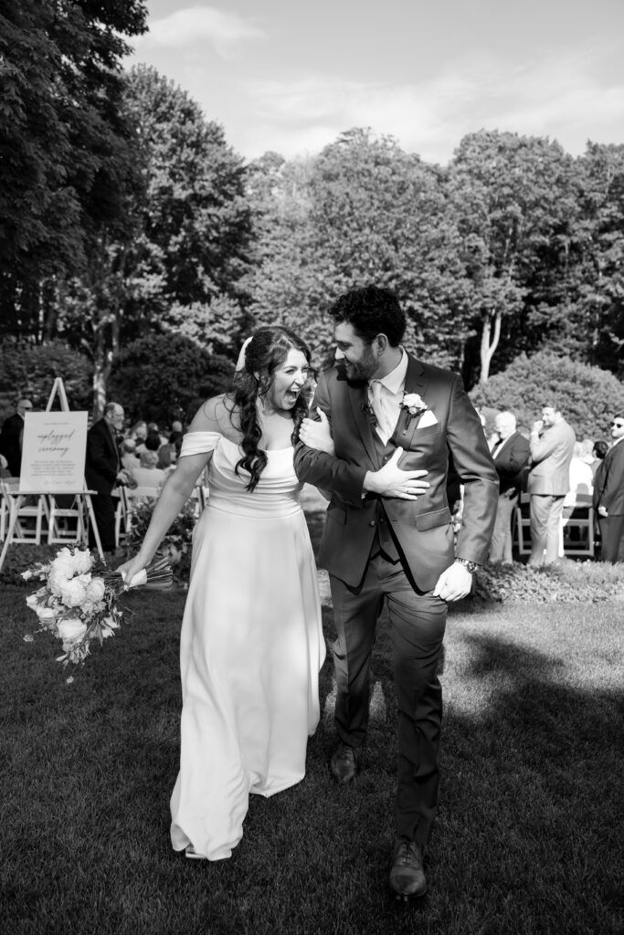 Smith Farm Gardens Wedding | Sincerely Liz Photography Documentary and Editorial Wedding Photography | Bride and groom walk joyfully arm-in-arm on a grassy path, surrounded by wedding guests. Trees and a bright sky form the backdrop. Black and white photo.