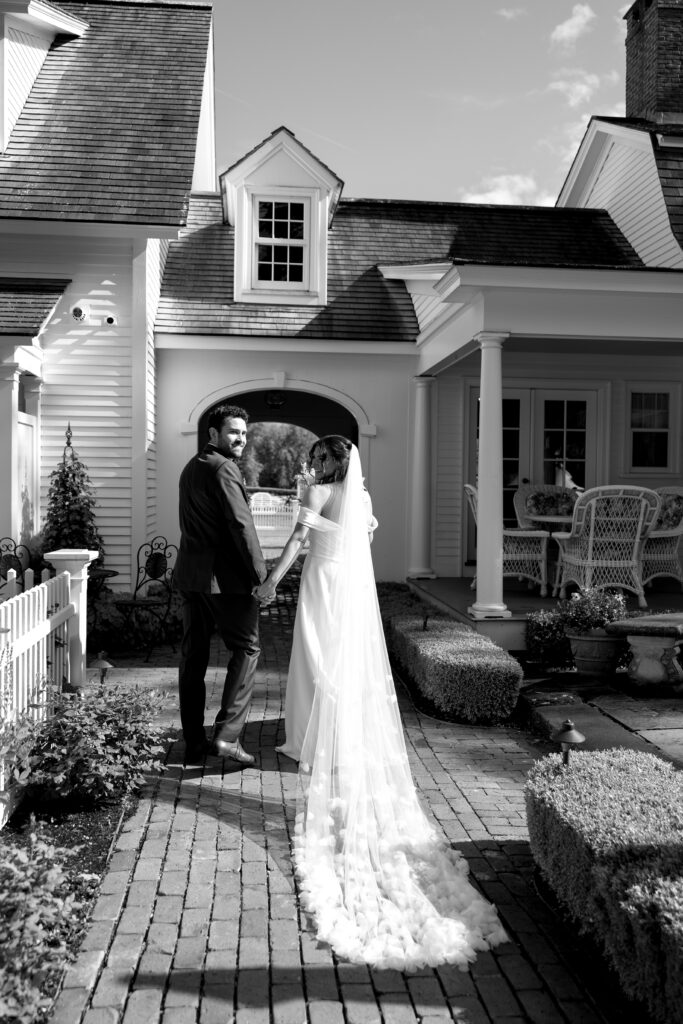In a black-and-white portrait, the couple walks hand in hand along a brick path outside a white farmhouse, reflecting the timeless charm of Smith Farm Gardens.