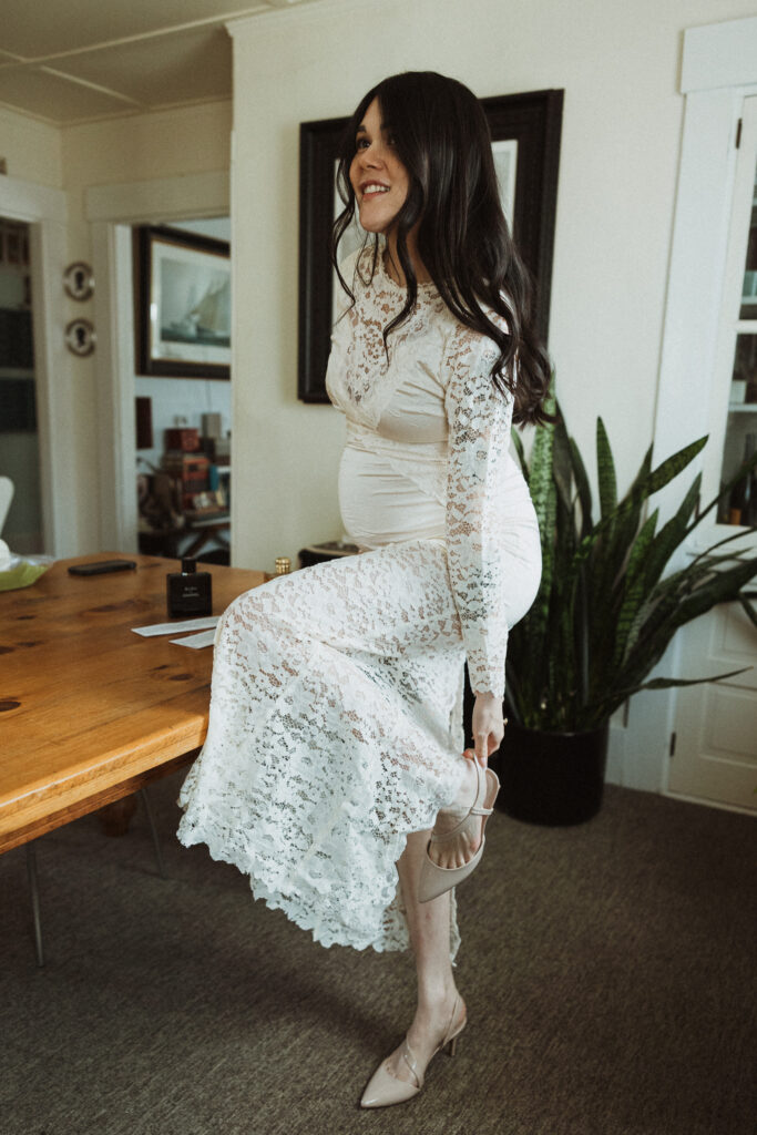 Boston Public Garden | Sincerely Liz Photography Documentary and Editorial Wedding Photography | Pregnant woman in a long white lace dress smiling, putting on beige heels in a cozy room with a wooden table and a tall houseplant nearby.