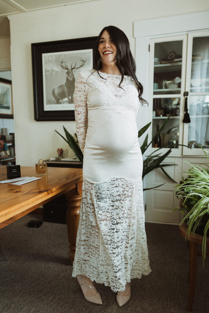 Boston Public Garden | Sincerely Liz Photography Documentary and Editorial Wedding Photography | Pregnant woman in a white lace dress stands smiling in a cozy room. Background features a wooden table, potted plants, and framed deer art.