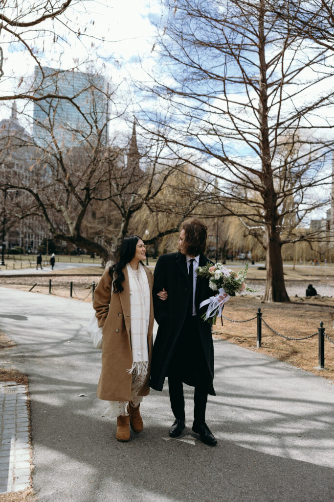 Boston Public Garden | Sincerely Liz Photography Documentary and Editorial Wedding Photography | Couple walking arm-in-arm in a park, smiling at each other. The man holds a bouquet, both in winter coats. Leafless trees and city buildings in the background.