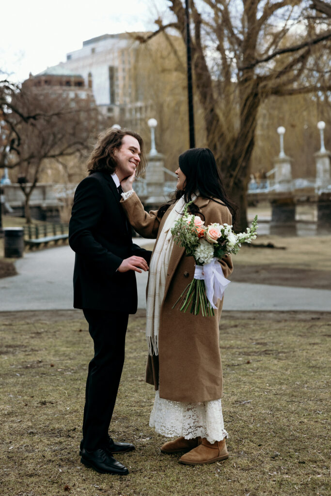 Boston Public Garden | Sincerely Liz Photography Documentary and Editorial Wedding Photography | A couple in formal attire shares a joyful moment in a park. The woman, holding a bouquet, gently touches the man's face. Bare trees and cityscape in the background.