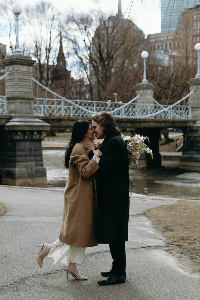 Boston Public Garden | Sincerely Liz Photography Documentary and Editorial Wedding Photography | A couple embraces affectionately on a park path near an ornate bridge. The woman wears a beige coat over a white dress, holding a bouquet. The scene is romantic and serene.