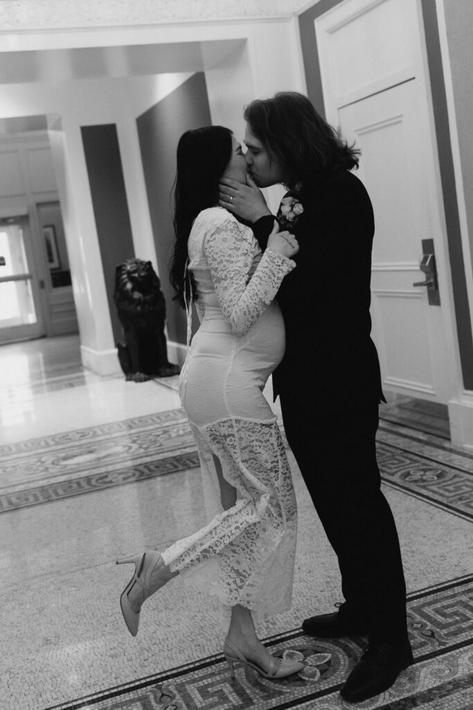 Boston Public Garden | Sincerely Liz Photography Documentary and Editorial Wedding Photography | A couple shares a tender kiss in a hallway. The woman, in a lace dress, lifts one leg playfully. The man, in a suit, holds her closely. Romantic and joyful atmosphere.