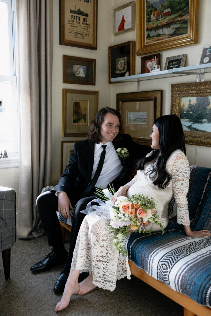 Boston Public Garden | Sincerely Liz Photography Documentary and Editorial Wedding Photography | A couple sits on a couch in a warmly lit room filled with framed artwork. The woman in a white lace dress holds a bouquet of roses, smiling at the man in a suit. The atmosphere is intimate and joyful.