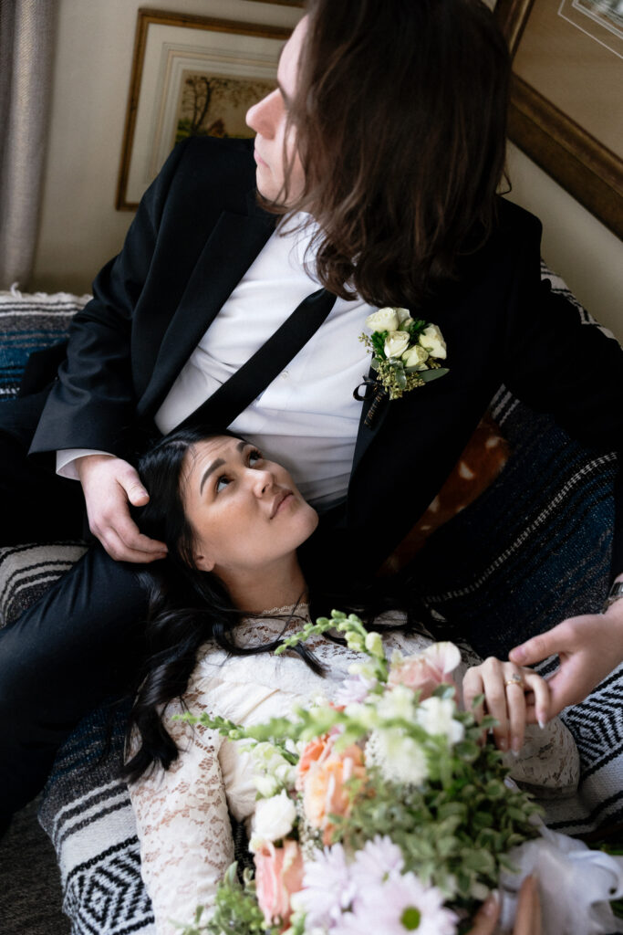 Boston Public Garden | Sincerely Liz Photography Documentary and Editorial Wedding Photography | A couple in wedding attire share a tender moment indoors. The woman rests her head on the man's lap, holding a bouquet, and they gaze at each other affectionately.