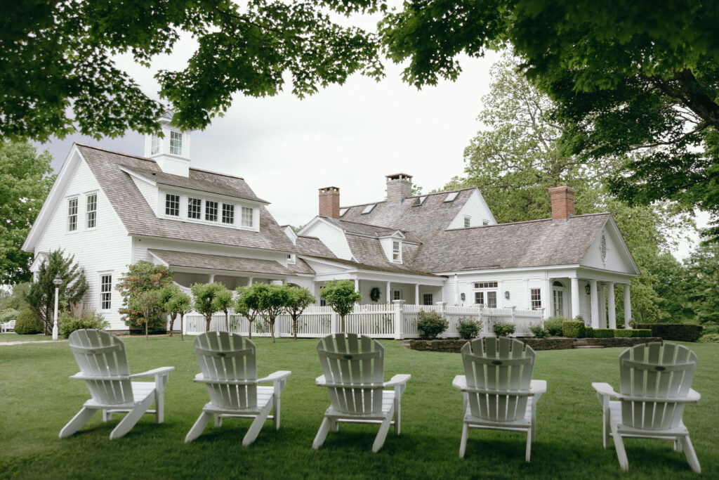 Smith Farm Gardens Wedding | Sincerely Liz Photography Documentary and Editorial Wedding Photography | Charming white house with a shingled roof, white picket fence, and lush lawn. Four Adirondack chairs face the house under leafy tree branches. Serene atmosphere.