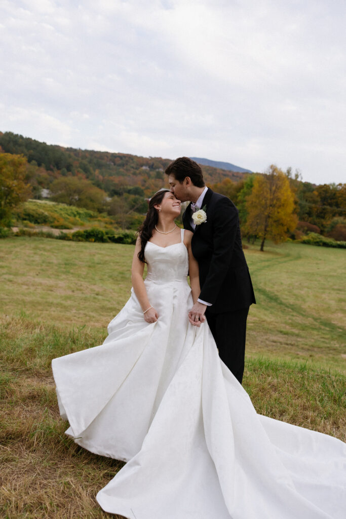 A bride and groom stand together in an open field with rolling hills in the distance, emphasizing the natural beauty of mountain wedding venues in Vermont.