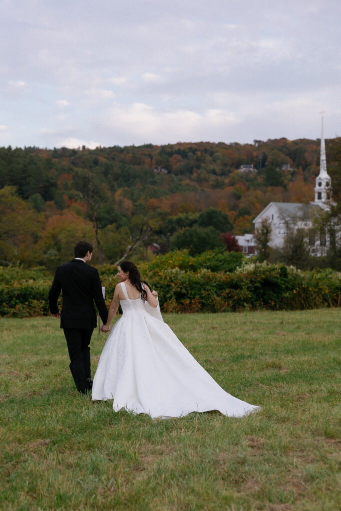 Wedding Photographers in Vermont | Sincerely Liz Photography Documentary and Editorial Wedding Photography | A bride and groom walk hand in hand across a grassy field, dressed in a white wedding gown and black suit. A scenic church and autumn trees are in the background, conveying a romantic and serene atmosphere.