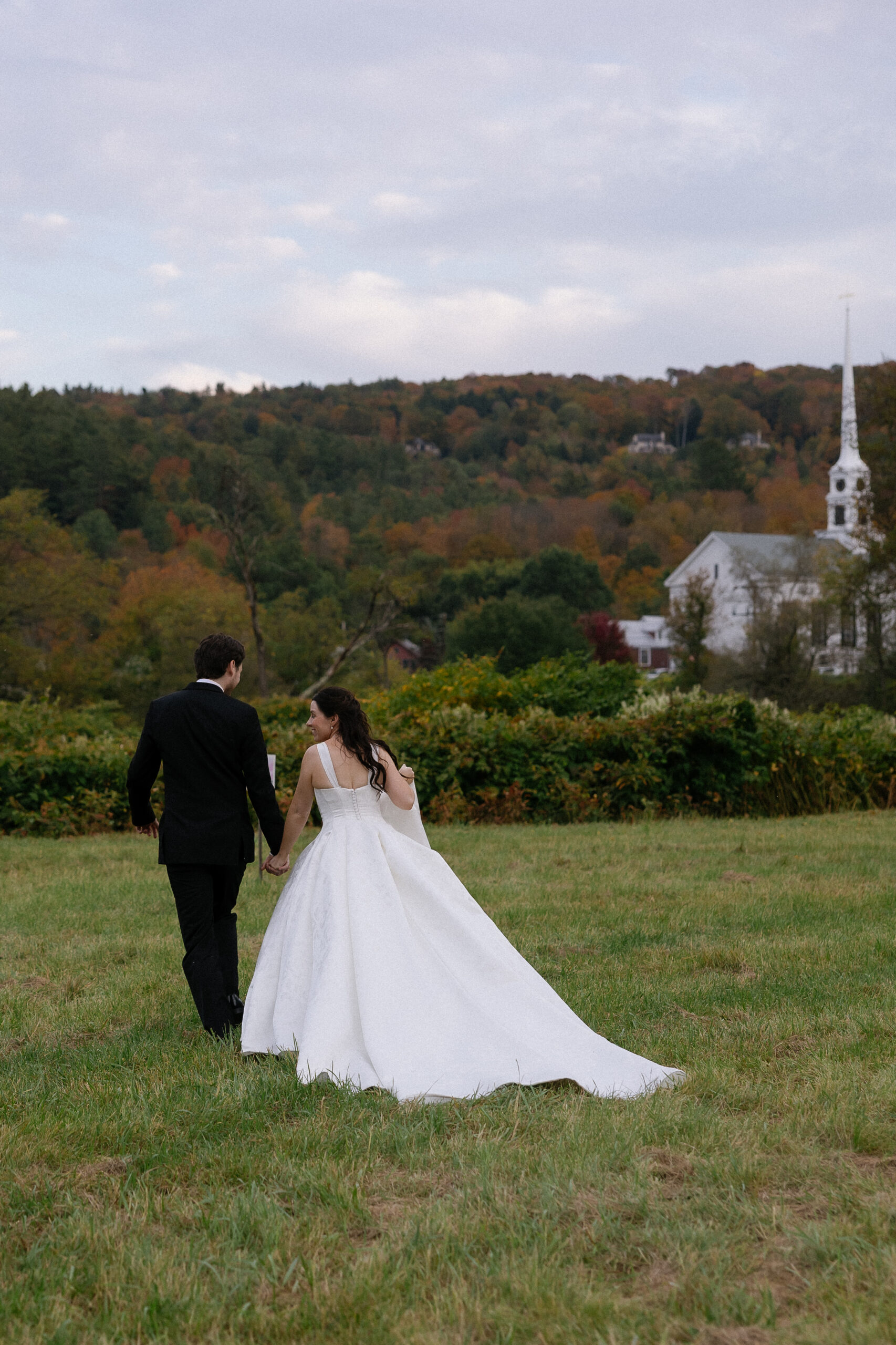 Wedding Photographer in Vermont | Sincerely Liz Photography Documentary and Editorial Wedding Photography | A couple in wedding attire walks hand in hand across a grassy field. The bride wears a white gown; the groom is in a black suit. Autumn trees fill the background.