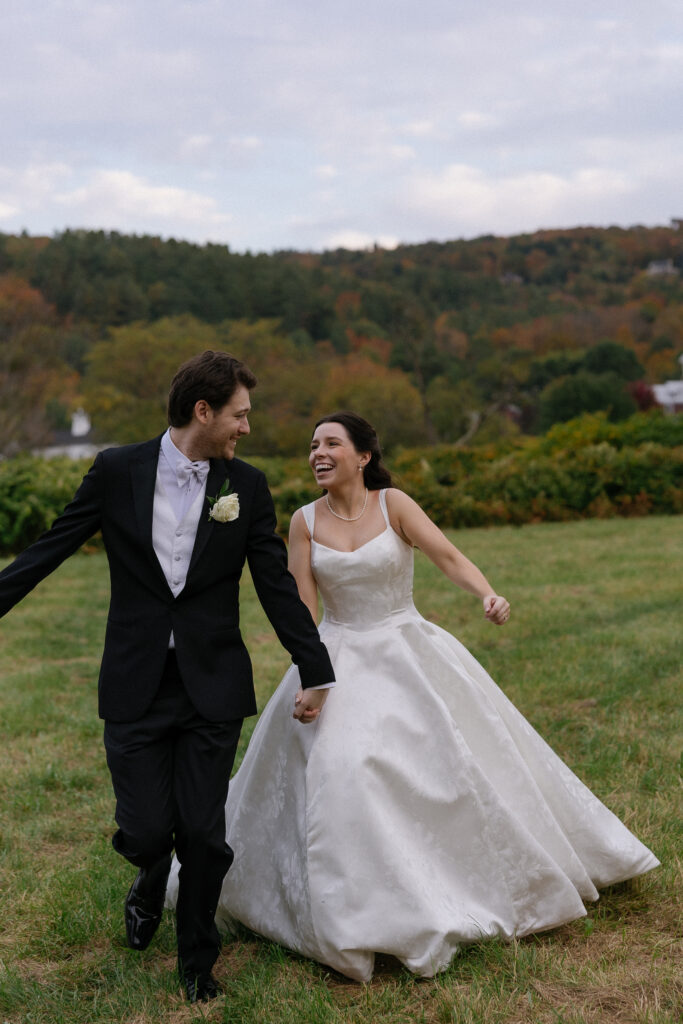 Wedding Photographer in Vermont | Sincerely Liz Photography Documentary and Editorial Wedding Photography | A joyous couple runs hand-in-hand across a grassy field, the bride in a flowing white gown and the groom in a black suit. The background shows autumn trees under a cloudy sky.
