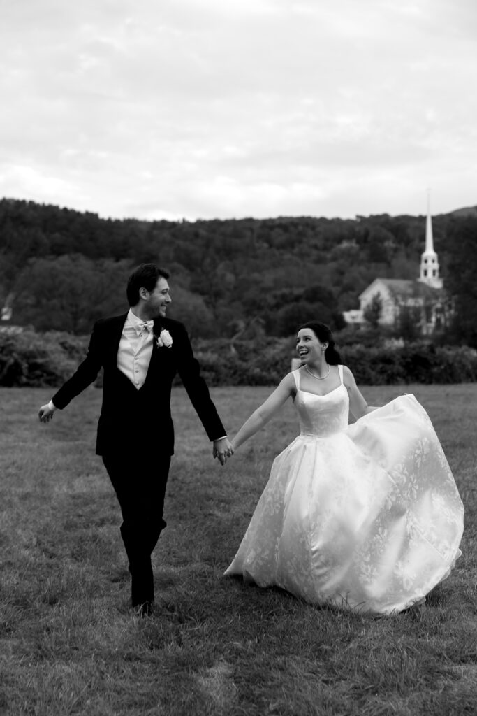 In black and white, the couple runs hand in hand across an open field with hills behind them, capturing the joy of a wedding in Vermont.