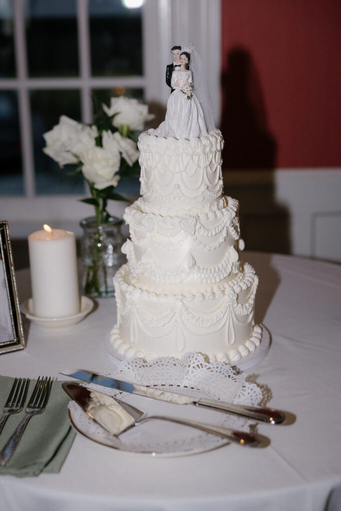 Wedding Photographer VT | Sincerely Liz Photography Documentary and Editorial Wedding Photography | Elegant three-tier white wedding cake adorned with ornate icing patterns and a bride and groom topper. Nearby, a lit candle, white roses, and cutlery set create a romantic ambiance.