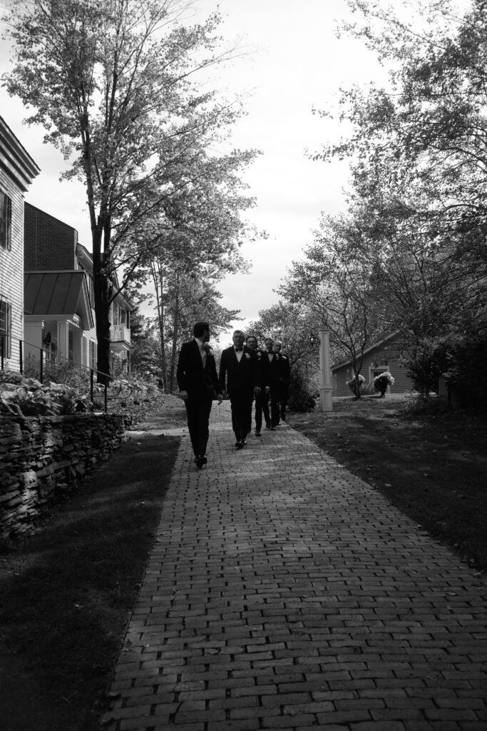 Guests walk along a shaded brick path between stone walls and trees in a black and white candid, a scene that reflects the atmosphere of best Southern Vermont wedding venues.