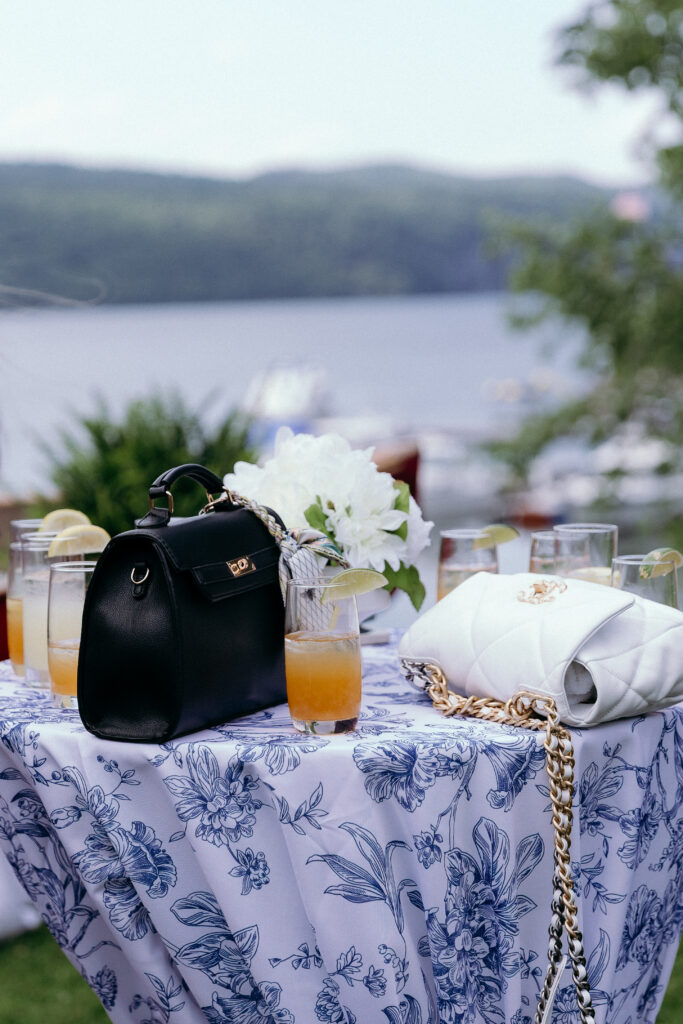Wedding Photographers in Vermont | Sincerely Liz Photography Documentary and Editorial Wedding Photography | Elegant outdoor setup with a floral-patterned tablecloth, black and white purses, cocktails, and white flowers. A serene lake and greenery are in the background.