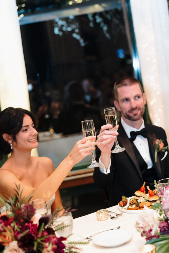 The couple raises champagne glasses during a candlelit reception, surrounded by warm tones and floral details that define elegant wedding venues in Vermont.