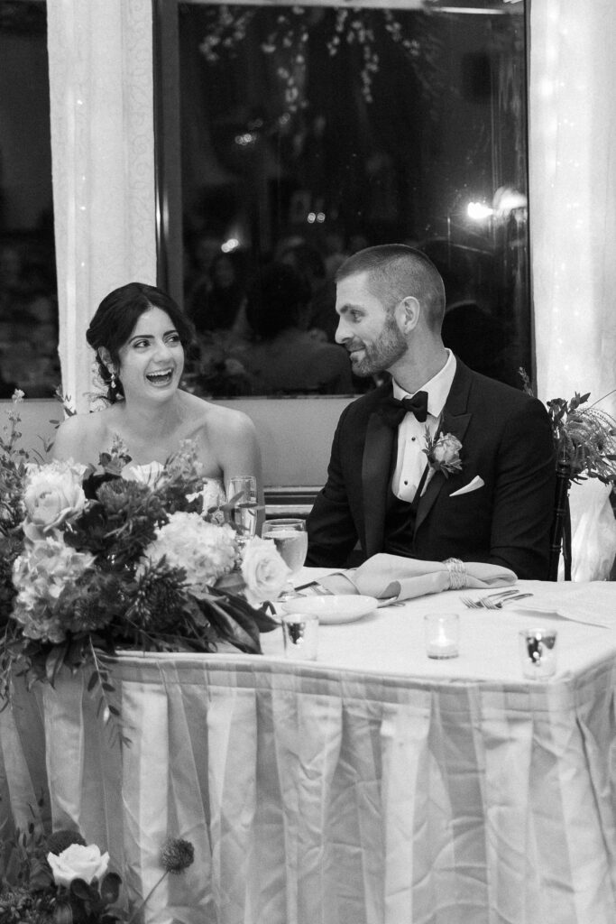 Wedding Photographer in Vermont | Sincerely Liz Photography Documentary and Editorial Wedding Photography | A smiling bride and groom sit at a table adorned with flowers and candles. The groom wears a suit, the bride a strapless dress. The moment is joyful and intimate.