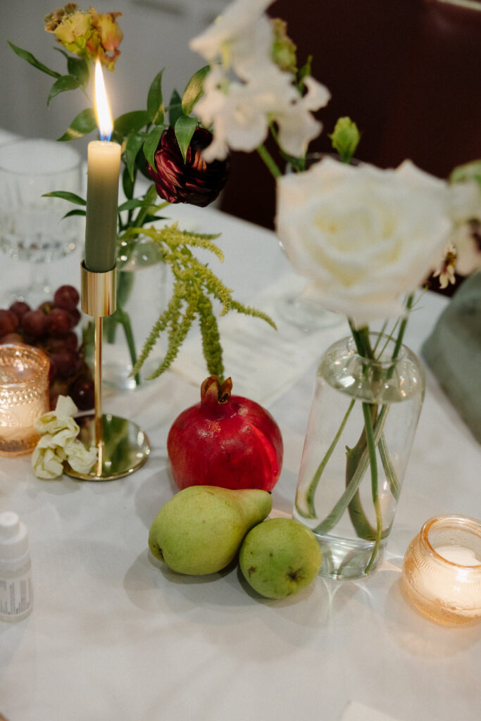 Wedding Photographer VT | Sincerely Liz Photography Documentary and Editorial Wedding Photography | A cozy table setting with a lit green candle, flowers in a vase, and fruits including a pomegranate and pears on a white cloth, creating an elegant, warm ambiance.