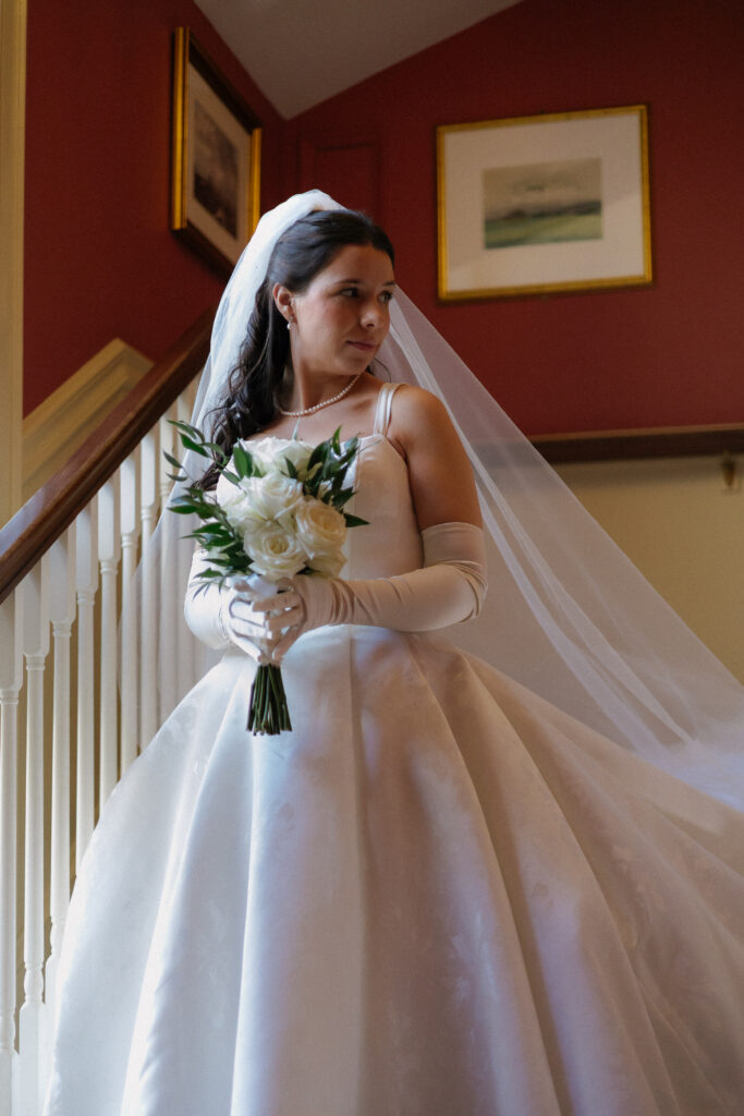 A bride stands on a staircase holding a bouquet, her veil draping softly behind her against warm-toned walls, capturing a classic interior moment often seen at the most stunning wedding venues in Vermont.