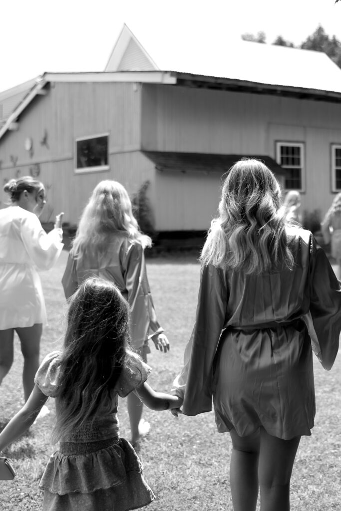 Wedding Photographer in Vermont | Sincerely Liz Photography Documentary and Editorial Wedding Photography | Black and white image of a group of women and a child walking towards a rustic house. They're in casual attire, holding hands, conveying a serene, familial atmosphere.