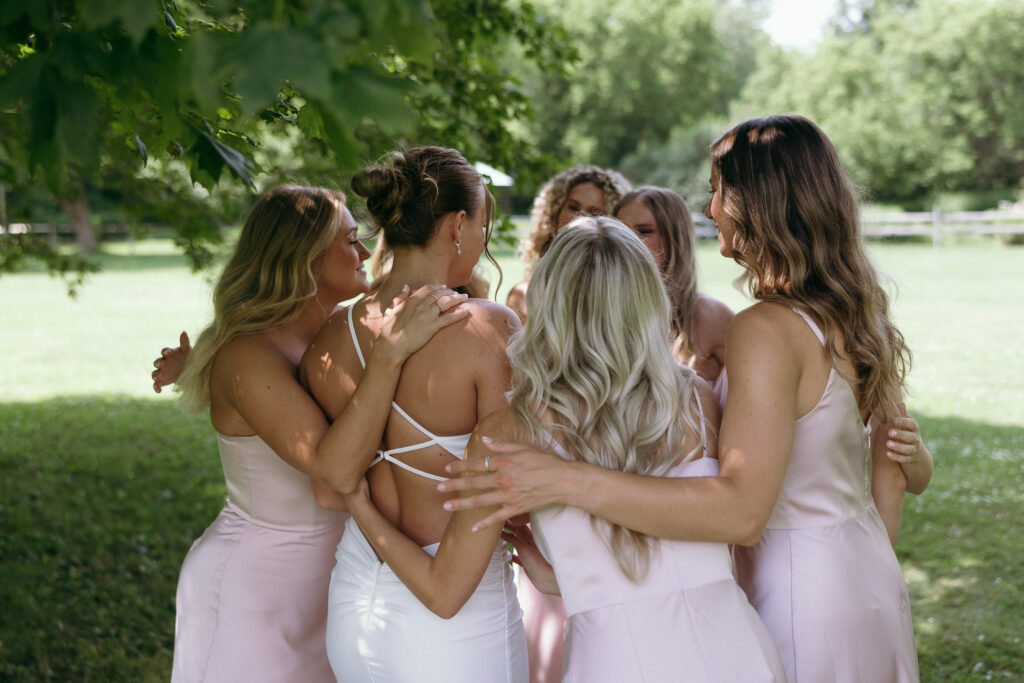 Wedding Photographer VT | Sincerely Liz Photography Documentary and Editorial Wedding Photography | A group of women in elegant pastel dresses embrace outdoors under lush trees, conveying joy and friendship in a sunlit, natural setting.