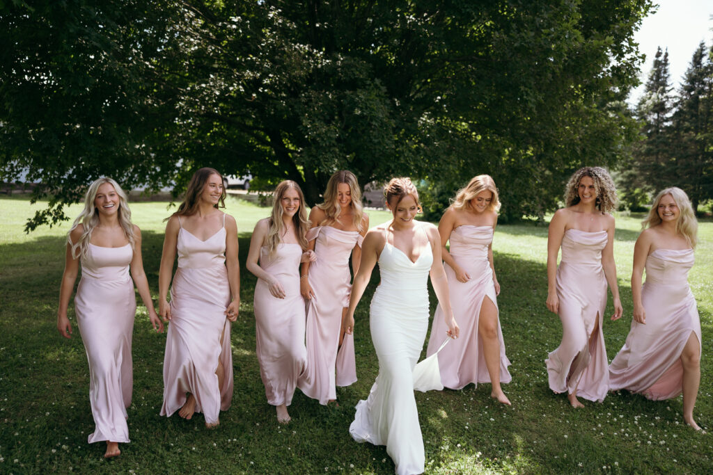Wedding Photographers in Vermont | Sincerely Liz Photography Documentary and Editorial Wedding Photography | A bride in a white dress and four bridesmaids in pink dresses walk on grass under sunlight, smiling, with lush green trees in the background.